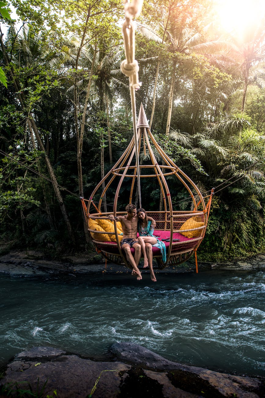 man and woman sitting on big rattan hammock above river surrounded with green trees